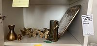 Shelf holding brass items including two vases, butterfly napkin rings, a tray leaning in the back, and a small rounded brass item.