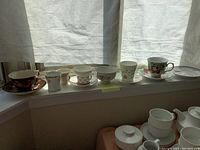 Window ledge with seven cups and saucers or cups alone arranged in a row, under natural light with sheer curtains behind them