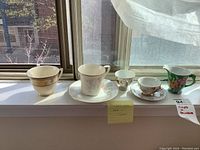 Five items displayed on windowsill in sunlight: Mikasa cup without saucer, Noritake cup and saucer, hand-painted miniature cup, and floral creamer