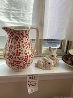 Photo showing two white Goebel rabbit figurines beside a Portuguese handmade pitcher with red flower pattern