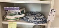 Shelf with four blue and white folded cotton placemats, several unopened packs of decorative napkins, and stacked paper plates.