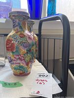 Photo shows one porcelain vase in front with detailed colorful floral and fruit decorations and one vase partially visible behind it on a white cloth.