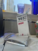 Square clear glass vase with a label showing lot details, placed on a table with other decor items in the background.