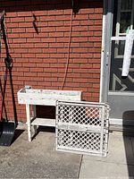 White painted wooden planter box with lower shelf and extendable white plastic baby gate folded and placed side by side on a concrete ground against a brick wall and door.