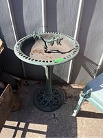 Full view of the metal bird bath showing pedestal base and bowl with decorative center branch and open hole rim design. Weathered, with debris in bowl.