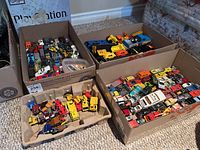 Photo showing multiple boxes containing a mix of Hot Wheels cars, trucks, and toy vehicles packed tightly on carpeted floor next to a couch.