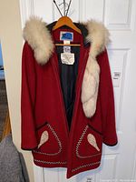 Full front view of red women's jacket on hanger, showing fur trim hood, patch pockets with beige stitched decoration, and zipper front.