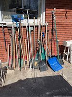 Wide shot showing all garden tools lined up against brick wall including brooms, shovels, rakes, hedge trimmers, metal storage rack, and plastic snow shovels.