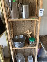 Shelf displaying two metal pitchers with rust, two metal strainers, glass peanut butter jar, glass barrel-shaped vase, and a clear glass decorative vase. Shelf is rattan and not included in lot.