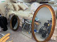 Photo showing three mirrors stacked vertically against a bed frame: two oval mirrors with ornate gold-colored frames and one rectangular mirror with a simple wooden frame.