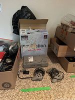 Sony PlayStation console with original box, two DualShock controllers, memory card on carpeted floor surrounded by boxes