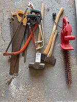 Overview photo showing all saws, axes, and red electric hedge trimmer together on concrete floor.