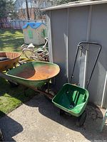 View of a rusted metal wheelbarrow with green frame next to a green Scotts seed spreader with black wheels beside a metal wall outdoors on a paved surface.