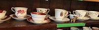 Full view of the teacups and saucers on wooden shelving, showing various patterns and brands