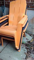Stacked plastic Muskoka chairs showing three orange chairs and one black chair underneath. Chair material is plastic with slatted seat and backrest design and wide armrests.