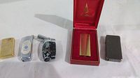 Photo shows five lighters in a row, including gold Zippo lighter, Forhu flameless lighter, Flamecrest Dom-33 gold lighter in red box, silver VW logo lighter, and black camo lighter.