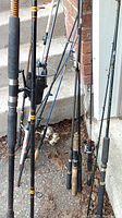 Photo showing seven fishing rods and several reels leaning against a wall on outdoor surface.
