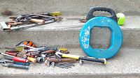 Mixed hand tools including hammer, screwdrivers, pliers, wrench, spool, and tape measure on concrete steps. Visible wear and rust.