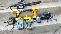 Power tools including two DeWalt cordless drills, a Craftsman electric chainsaw, batteries, and chargers laid out on concrete steps.