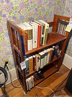 Front view of folding wooden bookshelf with books on shelves against floral wallpaper.