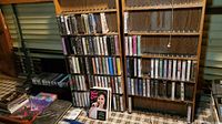 Wooden shelves filled with multiple rows of audio cassette tapes in plastic cases, some with handwritten labels, next to stacks of CDs and loose CD cases on a patterned floor.