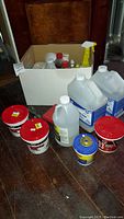Photo showing various home improvement and cleaning containers including paint thinner jugs, vinegar, red paint cans, and a box of assorted bottles.