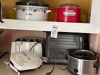 Shelf showing multiple kitchen appliances including crockpot, silver slow cooker, white toaster, rotisserie oven and red ice cream maker.