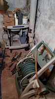 Photo showing the garden hose wound on a light green plastic spool with handle and wheels, alongside various garden tools including shears and cutters piled on the floor.