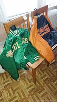 Two satin sports jackets hanging on wooden chairs; Celtics green jacket and Syracuse Orangemen orange/navy jacket.