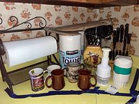 Photo of kitchen items including paper towel holder, paper towels, mugs, cutlery knives, utensil holders, and food prep tools arranged against a floral wallpaper.