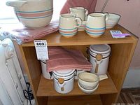 Side view of wood shelving unit holding McCoy stoneware items including bowls, pitcher, creamers, and ceramic canisters with wooden spoons. Pink kitchen towels folded in shelf.