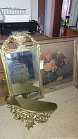 Photo showing full view of gold-toned mirror, matching shelf below it with scrollwork, and framed floral print leaning behind