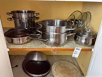 Shelf with various stainless steel pots and pans including large and medium pots, small pot with lid, and frying pan. Multiple glass lids are stacked on the right side. Also shows round wooden/bamboo cutting board below shelf and wok pan with wooden handle.