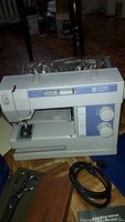 Front and right side view of white and light blue Viking Husqvarna LR15372 sewing machine with control dials and presser foot visible. Gingher shears and operating manual partially visible below machine.