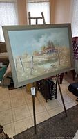Signed landscape painting on a brass easel, showing frontal view of the artwork and easel setup on floor indoors.