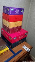 Stacked seven Trivial Pursuit game boxes in various colors representing different editions and sets from Vantage, placed on a wooden surface against a wall.