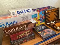 Photo showing vintage board games stacked, including Sequence, Electronic Sea Battle, Labyrinth, To Kill A Lawyer, Yahtzee, Play It By Ear, Save Butterfly, and a Backgammon set with open case.