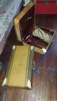 Photo showing two vintage suitcases on a dark wood floor, one open with brown satin-like lining and attached fabric bag inside, the other closed with yellow exterior and metal corners.