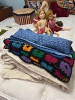Three crocheted afghans folded and stacked, with the bean bag and dolls in the background.