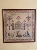 Framed antique needlepoint sampler on wall showcasing full artwork, floral border, and figures.