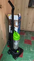 Front view of Bissell Easy Vac upright vacuum cleaner with green top and clear dust container, attachments on side, and box visible in background.