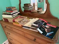 Photo showing assorted books including the Columbia Encyclopedia volumes stacked on a dresser with a mirror reflecting the room.
