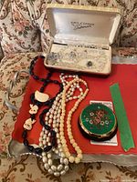 Overview of all items on tray showing several beaded necklaces, Sherman jewelry box with crystal necklace and earrings, and vintage makeup compact with floral enamel design.
