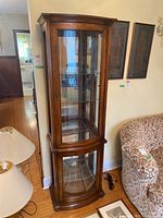 Full view of the tall wooden display cabinet with glass shelves and brass key locks, situated in a room next to furniture and a lamp.