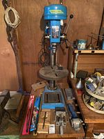 Front view of blue Brico SL-M4S drill press with base, worktable, and accessories at foot.