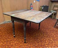 Front angled view of the antique wooden drop leaf farm table showing the open extended surface and turned legs with worn paint ends.