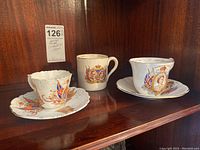 Three commemorative china items displayed on shelf showing the smaller Aynsley cup and saucer, larger Taylor and Kent cup and saucer, and the mug with royal portraits.