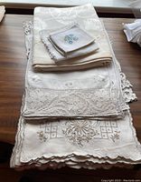 Stack of folded linens including embroidered napkins and several larger tablecloths with floral and lace detailing.