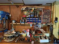 Full view of the workbench surface and various hand tools placed on it including hammers, pliers, and screwdrivers; drill press and shelves with containers and jars in background.