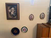 Wide view showing two petitpoint needlepoint oval frames, two small Limoges plates beneath, and a floral motif shadow box on the left side.